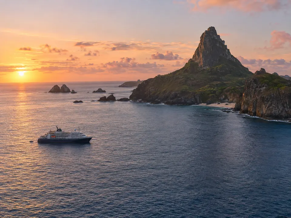 O Morro do Pico ao amanhecer, visto do mar, com um navio de expedição ancorado em primeiro plano.